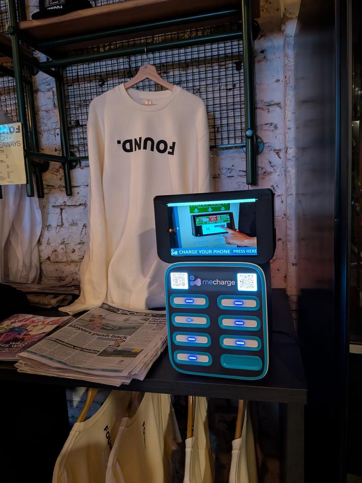 A phone charging station with a small screen displaying an ad, inside a gaming or entertainment center with a banner for Zero Latency Virtual Reality Arena in the background.