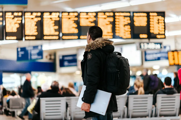 A man with a backpack and a laptop in hand at a busy airport terminal looking at an electronic flight information display board