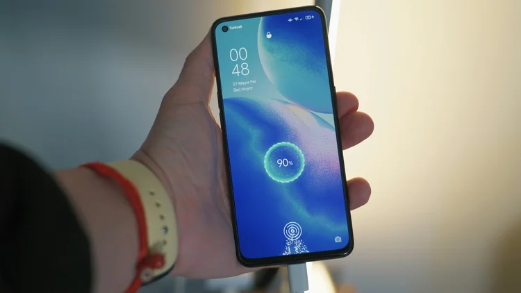 A person holding a smartphone with a charging screen showing 90% battery, time 00:48, and the date May 27, with a lock icon and a small profile icon at the top, and a charging symbol at the bottom.